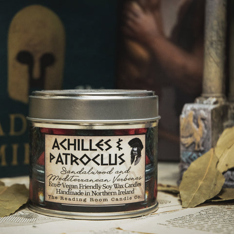 Soy Wax Candle labeled 'Achilles & Patroclus' on a surface with books and leaves in the background
