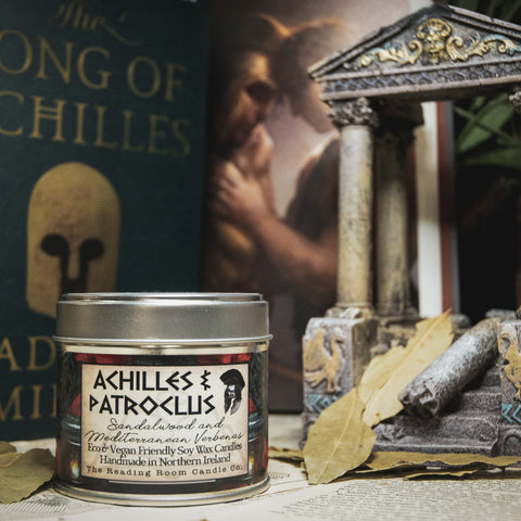 Soy Wax Candle labeled 'Achilles & Patroclus' on a surface with books and leaves in the background