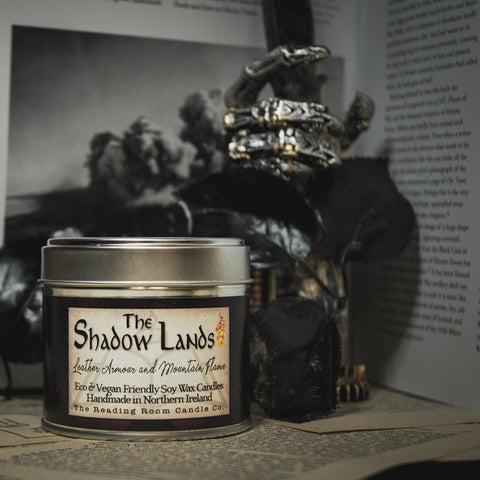 Soy Wax Candle tin labeled 'The Shadow Lands' on an open book with a dark, moody background.