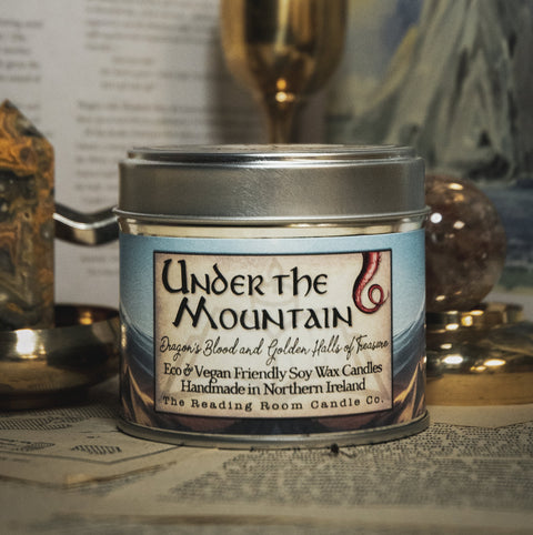 Candle jar labeled 'Under the Mountain' on a surface with books and decorative items in the background