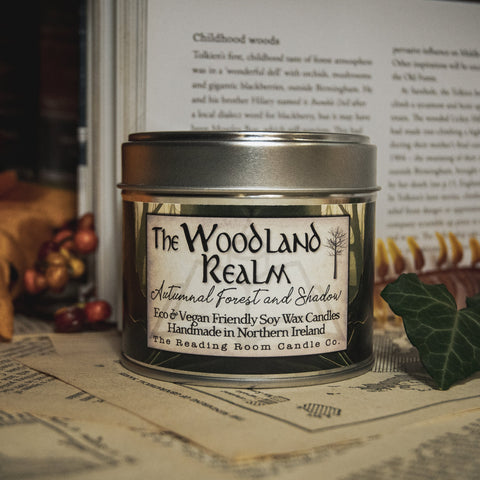 Soy Wax Candle labeled 'The Woodland Realm' on a surface with leaves and an open book in the background