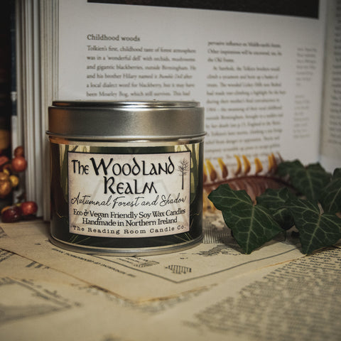 Soy Wax Candle labeled 'The Woodland Realm' on a surface with leaves and an open book in the background