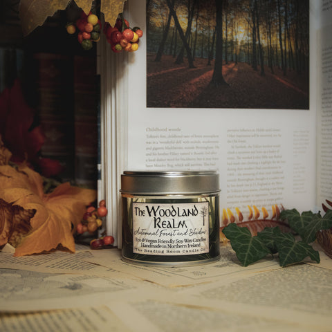 Soy Wax Candle labeled 'The Woodland Realm' on a surface with leaves and an open book in the background